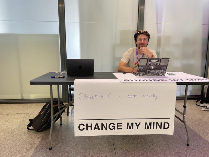 A desk with a large "Objective-C is great actually, CHANGE MY MIND" banner. Sitting at a desk is another Objective-C afficionado (disputed).