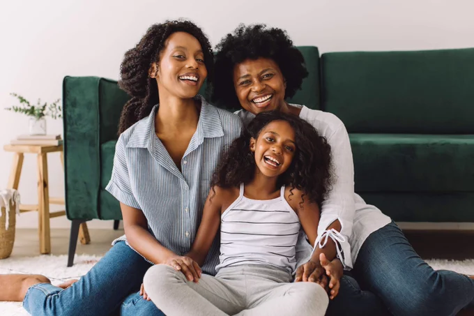 Three Black women, two adults and one child, smile together in their living room. A green couch is behind them.