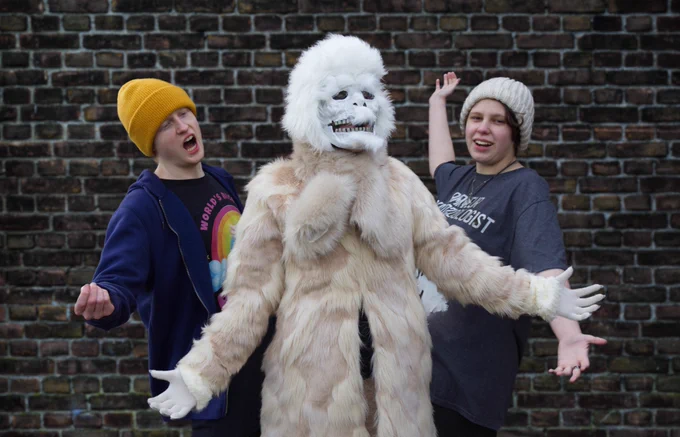 A still from the film "Have You Found It Yet?." A person dressed up as a yeti in a fur coat and a mask standing shrugging in front of a brick wall. Two people stand either side of the yeti with their arms stretched out in presenting mode.