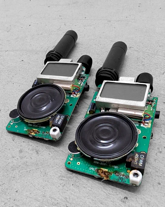 The image shows two identical electronic devices with exposed circuit boards. Each device has a small black speaker in the center, a square LCD screen above it, and a short, black antenna protruding from the top. The components are mounted on green printed circuit boards with visible wiring and connections. The devices appear to be in a partially assembled or disassembled state, possibly as part of communication equipment or small radios. The background is a plain, gray surface, likely a workbench or floor.