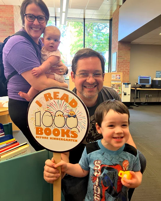 A mother, father, and two kids smiling at the camera. The child in front is holding a sign that says "I read 1000 books before kindergarten!"