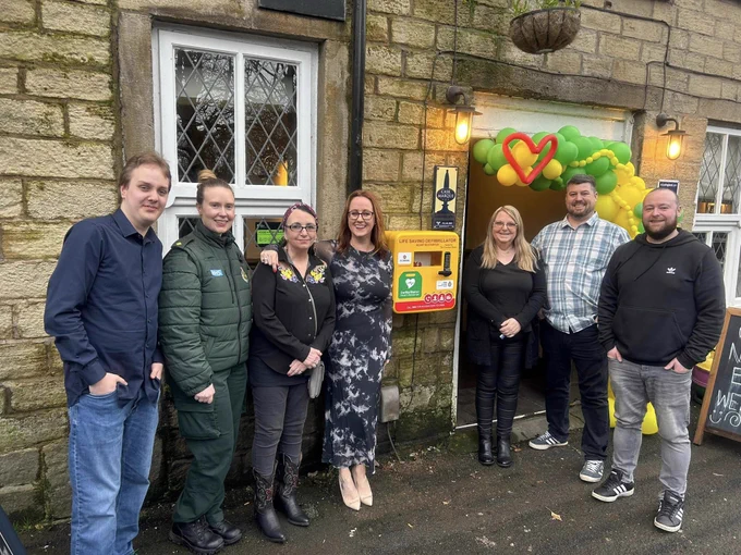Group of seven adults standing outside a stone building next to a defibrillator installation, with a decorative balloon artwork in the background.