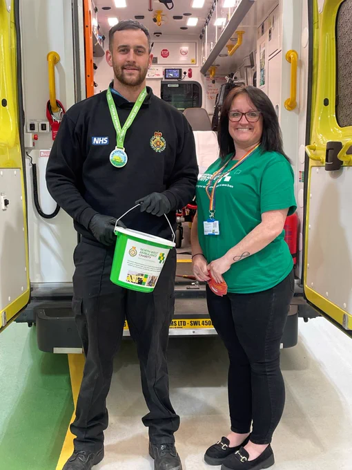 Two individuals from the NHS standing by an ambulance, one wearing a green NHS uniform and a medal, the other in a green shirt, holding a donation bucket.