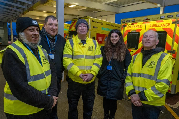 Group of five emergency service personnel wearing high-visibility vests and warm hats, standing in front of a North West Ambulance Service vehicle.