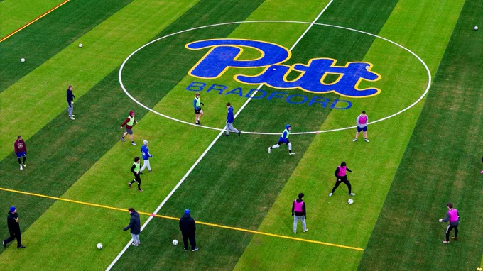 A group of people participating in a soccer training session on a field with 'Pitt Bradford' logo at the center.
