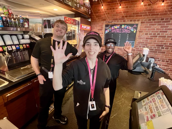Zach, Rebecca, and MC Dan wave and smile in Concessions.