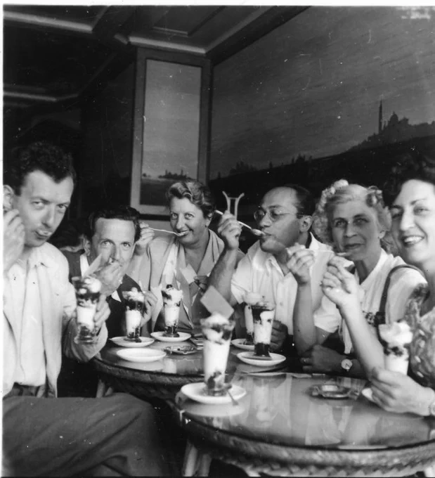 A black and white photo of three men and three women in early 1950s fashions, all gathered at two small round cafe tables eating ice-cream from tall glasses with long spoons.  (These are members of the English Opera Group on tour in Italy in the early 1950s: the composer Benjamin Britten and his partner the singer Peter Pears are on the left.)