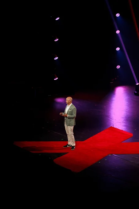 An individual presenting on a TEDx stage, standing on a large red "X" with stage lighting overhead.