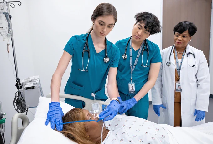 Two students in scrubs are examining a patient lying in a hospital bed, while another professional in a white coat observes. 