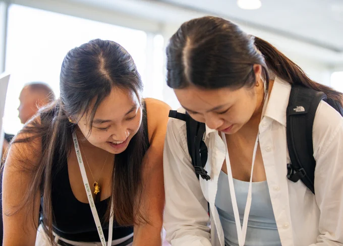 Two Rush University students, both wearing lanyards, are looking closely at a document together with smiles.