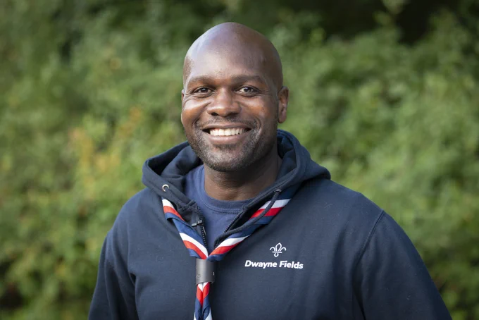 New UK Chief Scout Dwayne Fields smiling, wearing a Scouts necker and navy blue hoodie.