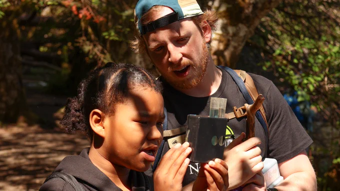 A young student and a NatureBridge educator look together at a handheld pH reader holding two test tubes filled with liquid, one clear and one blue.