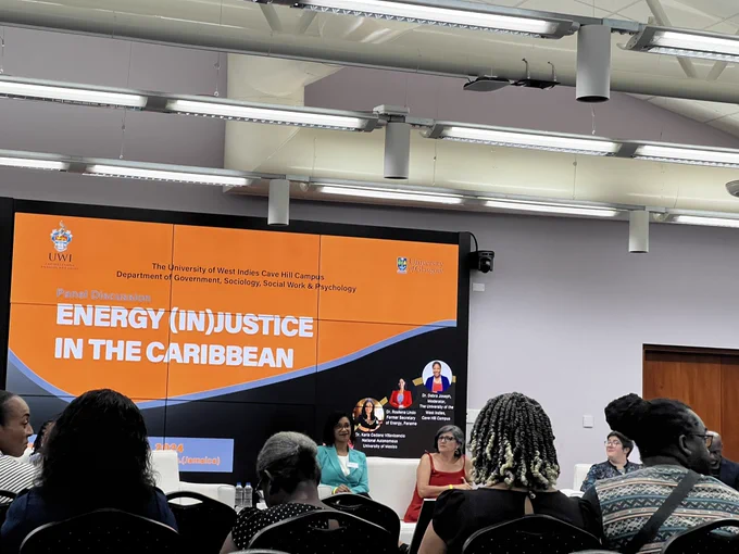 Photo of panel showing Rosilena lindo wearing a turquoise suit, Karla Cedano in a red dress, and Harriet Thomson wearing a floral dress. In the photo several rows of people sitting can be seen, as well as the screen that says “Energy (In)justice in the Caribbean” in white letters and orange background.