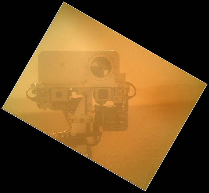 A reddish-and-gold-hued square shows the head of the Curiosity rover on Mars. This image was taken with its MAHLI instrument and the dust cover was on, which is why the image has the hazy tint. The rover's head is off-center in the image and it appears to be staring straight at the camera.