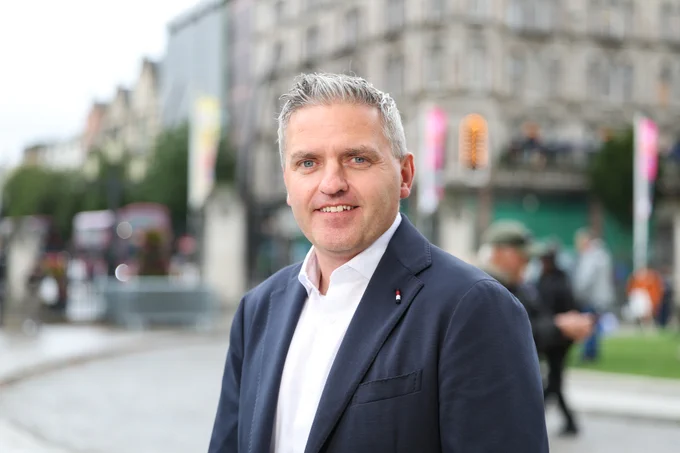 Dr Adrian Johnston standing front-centre with a white shirt and navy blazer amidst the backdrop of Belfast City Centre
