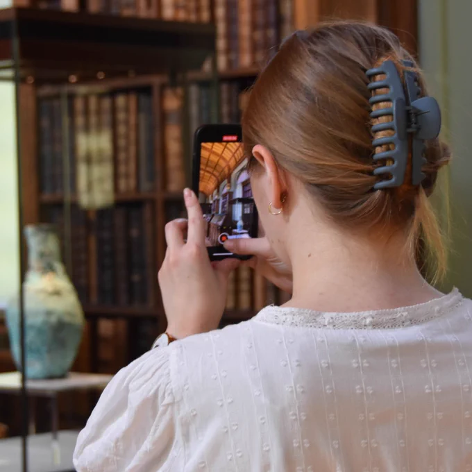 Person taking a photo with a smartphone inside a library with bookshelves and artefacts in the background.