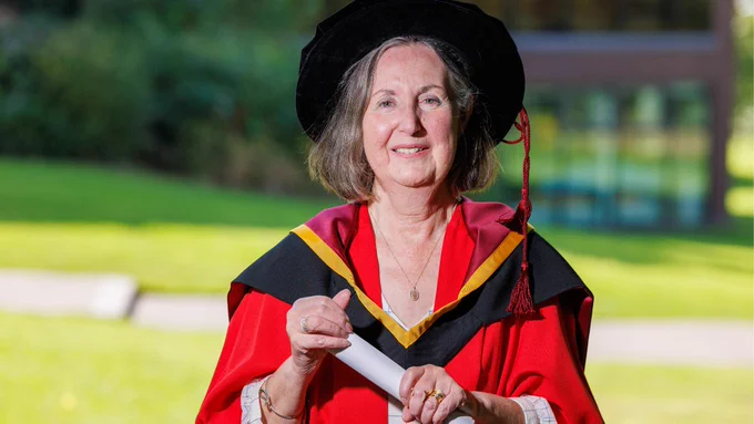 A photo of a woman from the elbows up, wearing red ceremonial robes and a black conferring cap, with a black and yellow sash on top and a white shirt underneath. She is smiling at the camera and holding a scroll in both hands, against a blurred background of green grass and a brown brick building. 