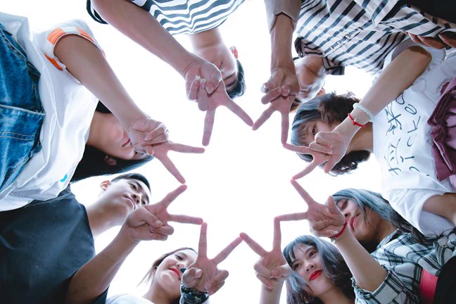 Eight young people stand in a circle, with a hand each in the center. They each have two fingers out, touching the fingers either side.