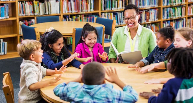 A group of younger children sit at a round table, with a teacher reading from a book. The children are all smiling and pulling faces, and are waving their hands above the table. 