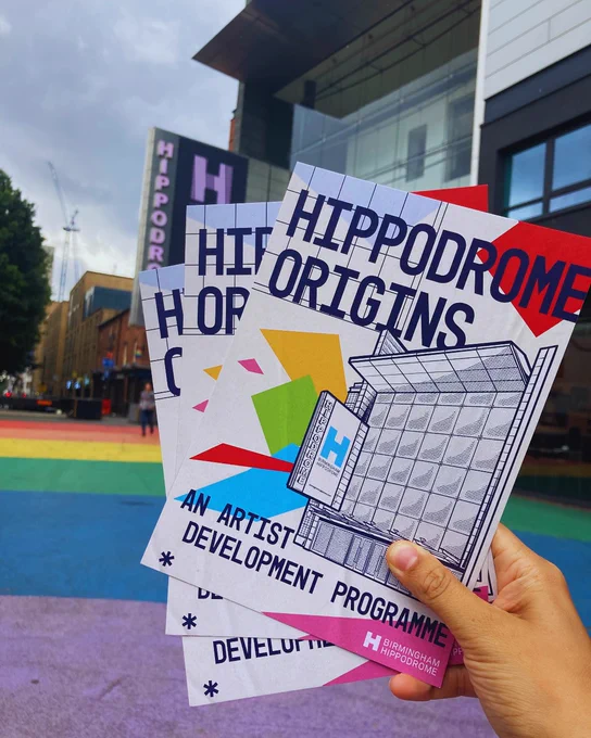 A hand holding colorful promotional flyers for "Hippodrome Origins - An Artist Development Programme" in front of the Hippodrome building.