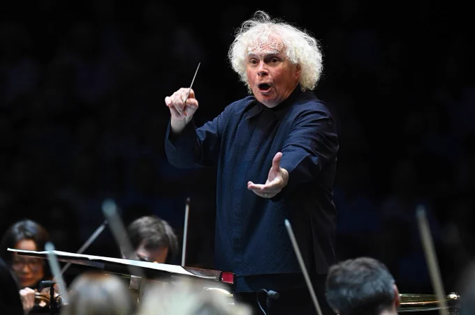 Sir Simon Rattle mid-movement as he conducts the Bavarian Radio Symphony Orchestra, one arm outstretched towards the musicians.
