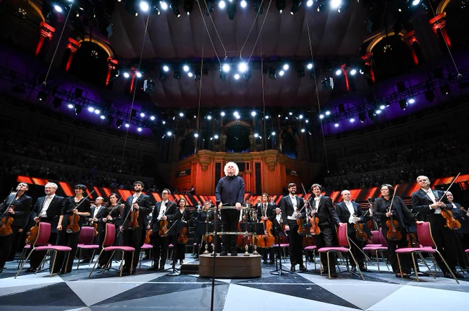 Sir Simon and the Bavarian Radio Symphony Orchestra stand on the Royal Albert Hall stage, facing the audience and the camera. 