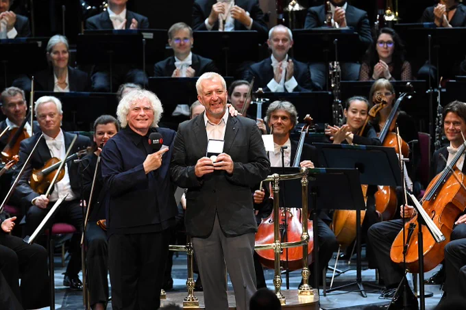 
Sir Simon Rattle stands on the Royal Albert Hall stage, smiling, holding a BBC Radio 3 branded microphone in his right hand, with his left arm around Thomas Adès. Thomas is also smiling and holding the Royal Philharmonic Society Gold Medal, which Sir Simon has just presented to him. Members of the Bavarian Radio Symphony Orchestra are seated behind them, smiling and applauding.