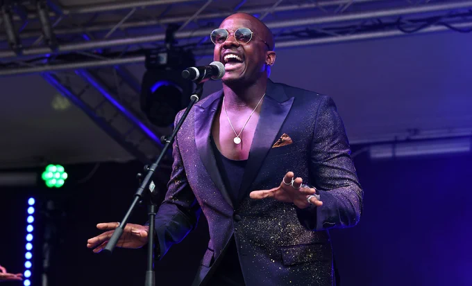 Performer singing passionately into a microphone on stage, wearing a sparkling dark blazer and sunglasses.