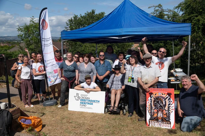 A group of people cheer happily under a blue tent, behind a large Heritage Fund sign. 