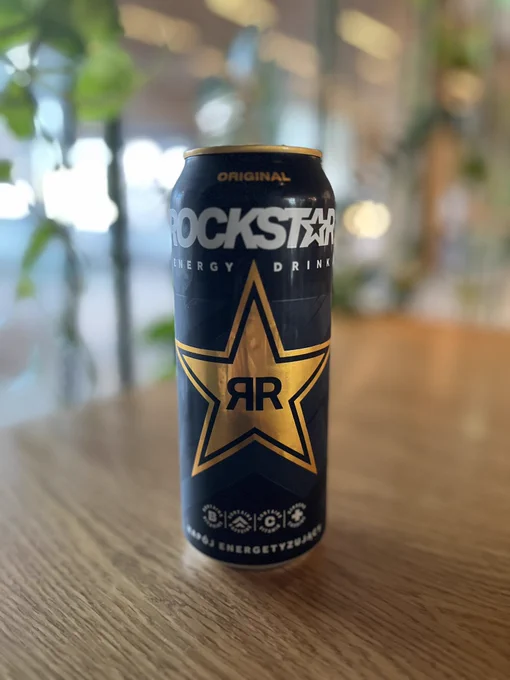 Rockstar drink in a tall black van with a gold star in the middle with various rockstar logos, on a wooden table with a blurred background.