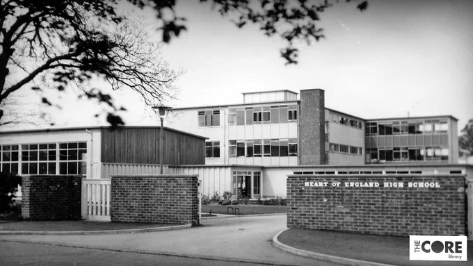 Black and white photograph of the exterior of a school building showing an entrance drive between two brick walls, looking towards a three-storey classroom block