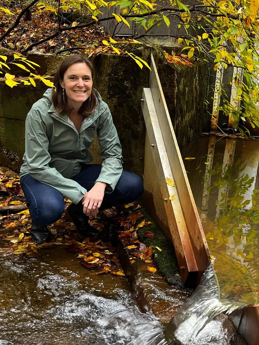 A female scientist wearing a green rain jacket crouches in a stream smiling at the camera, beside a V-shaped weir with water flowing over it.