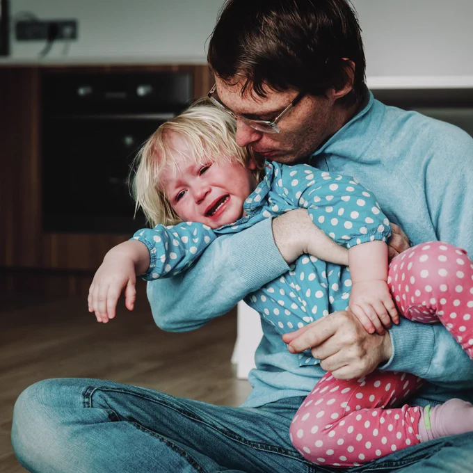 A father sitting on the floor with his crying toddler, trying to settle her
