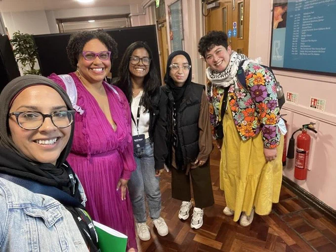 5 femme-presenting people in various shades of brown, two of which are wearing hijabs. Everyone is smiling.