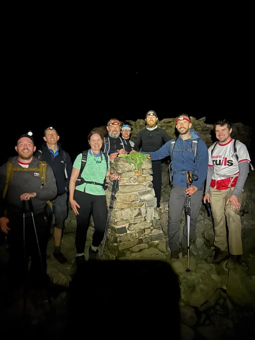 Image of a group of hikers at the summit stone of Scafell Pike. It is the middle of the night so the sky is dark and the camera flash is illuminating the group.