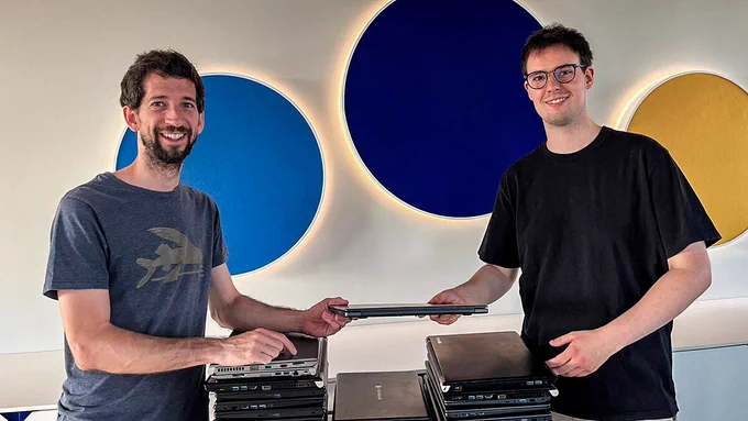 Im Büro der Firma Formycon AG stehen 2 Personen vor einer hellen Wand, darauf 3 große, hinterleuchtete Kreise in hell-, dunkelblau und gelb. Vom Betrachter aus rechts steht Patrick Stevens von Formycon, links Juan vom Labdoo Hub München. Beide halten einen Laptops rechts und links fest und schauen in die Kamera. Auf einem Tisch zwischen beiden liegen gestapelte Laptops.