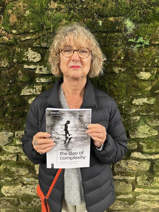 Jean Boulton stands in front of a mossy stone wall, smiling proudly and holding her new book up, showing the cover to the camera. The cover of the book features a grayscale watercolour paint illustration of what appears to be a silhouetted person walking along a watery landscape dappled with sunshine. The book title reads, 'The Dao of Complexity: Making Sense and Making Waves in Turbulent TImes'.