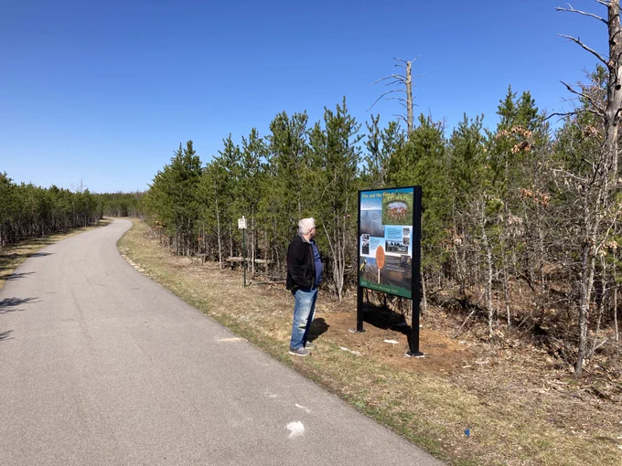 A man reading an interpretive sign along a paved trail surrounded by trees.