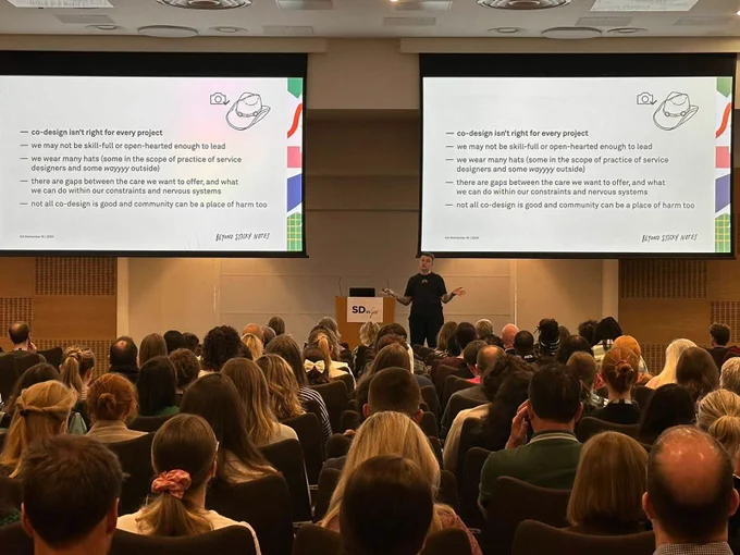 A person presenting at SDinGov logos on the screen behind them. The participants, seated in rows, attentively listens and takes notes. The presentation slide includes text about design rights for every project.