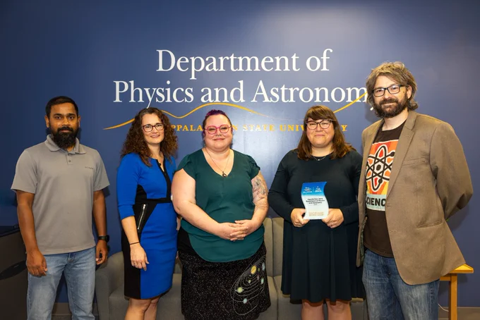 Five people stand in front of a blue wall, smiling. One person is holding a wavy object, which is the P/A SEA Change award.