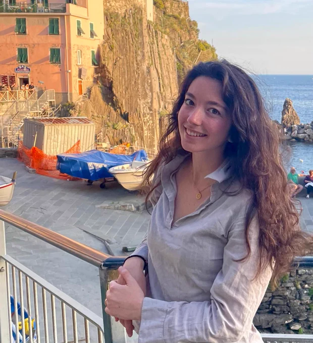 A picture from Martina Morchio posing in front of the sea, with what looks like a fishing port behind her, under sunset lightning.