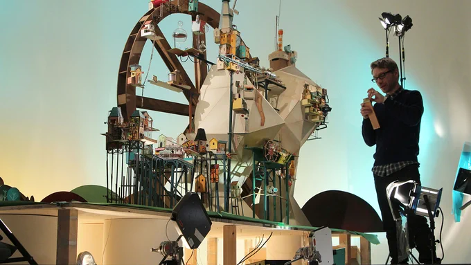 A person standing next to a large, intricate model, showing a large wheel with small houses around it. On the ground are studio lights and filming equipment