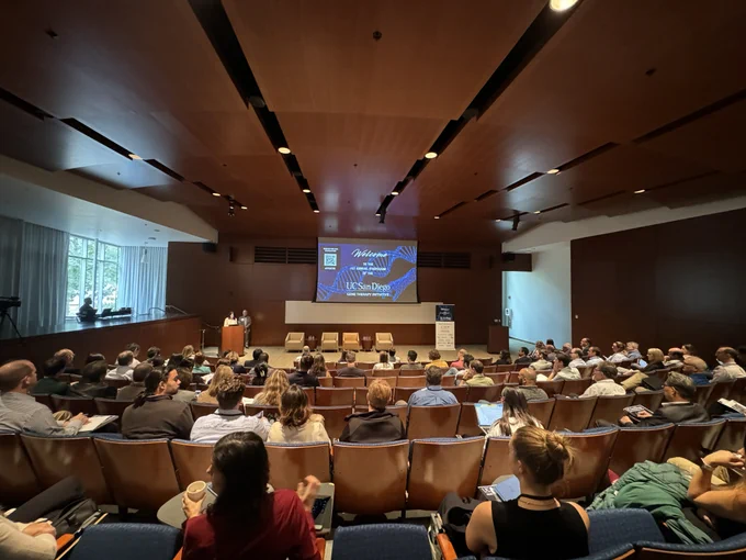 A large group of people in an auditorium watch two speakers at a podium