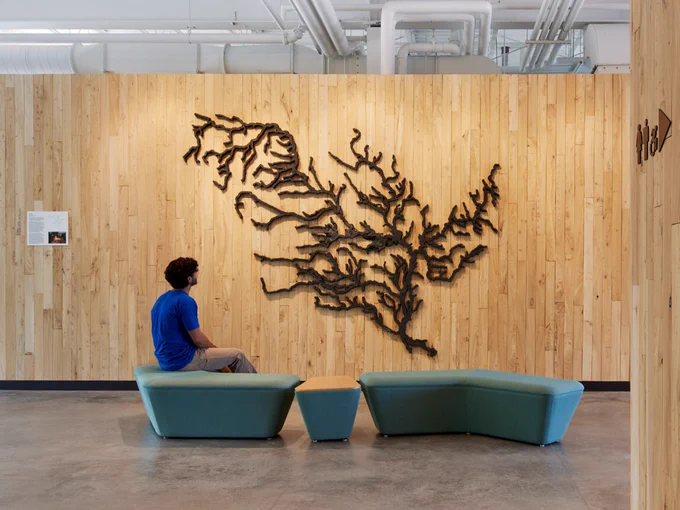 Mississippi River of Iron by Tamsie Ringler at CRWD’s Watershed Learning Center. Photo by Corey Gaffer. 
A person sitting on a blue bench, looking at a large iron sculpture of the Mississippi River watershed on a wood-paneled wall in a modern office.  