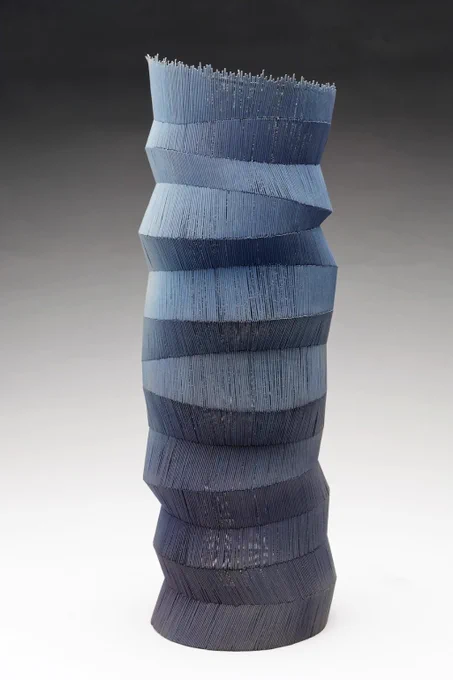 A tall blue vessel made of thin wires with a sectioned, tilting form stands against a white and grey background.