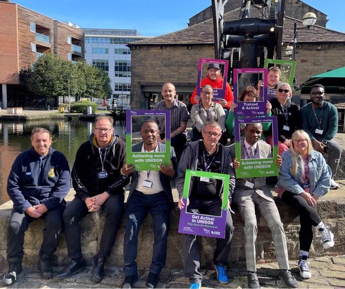 A group of people are sat in the sunshine smiling. Some of them are holding photo frames in UNISON colours, which say "Trained and active!", "Achieving more with UNISON" and "Get Active! in UNISON". They have completed a UNISON training course in Leeds.
