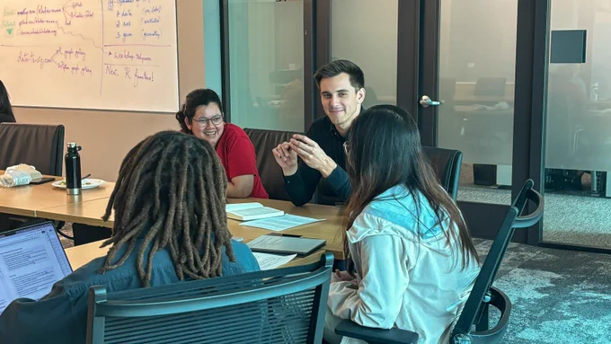 Four professionals are engaged in a collaborative discussion around a table in a modern office setting. The meeting room features a glass wall and a whiteboard partially covered with written notes.