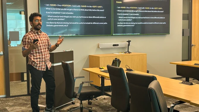 Person presenting in a conference room with a large table, chairs, and a presentation slide visible on a screen in the background.