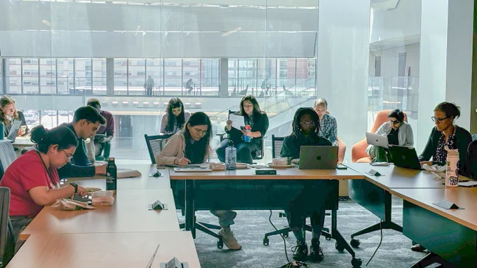 Group of professionals working on laptops and taking notes around a conference table in a brightly lit office space with large windows in the background.