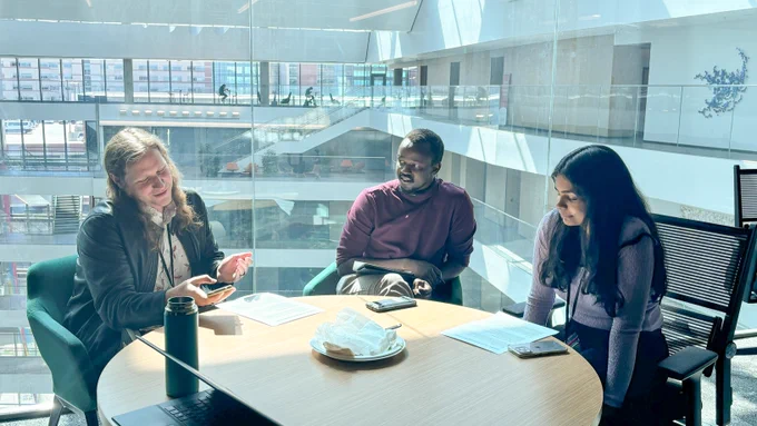 Three people seated around a glass table in a bright office setting, engaging in a conversation with documents and mobile phones on the table.
